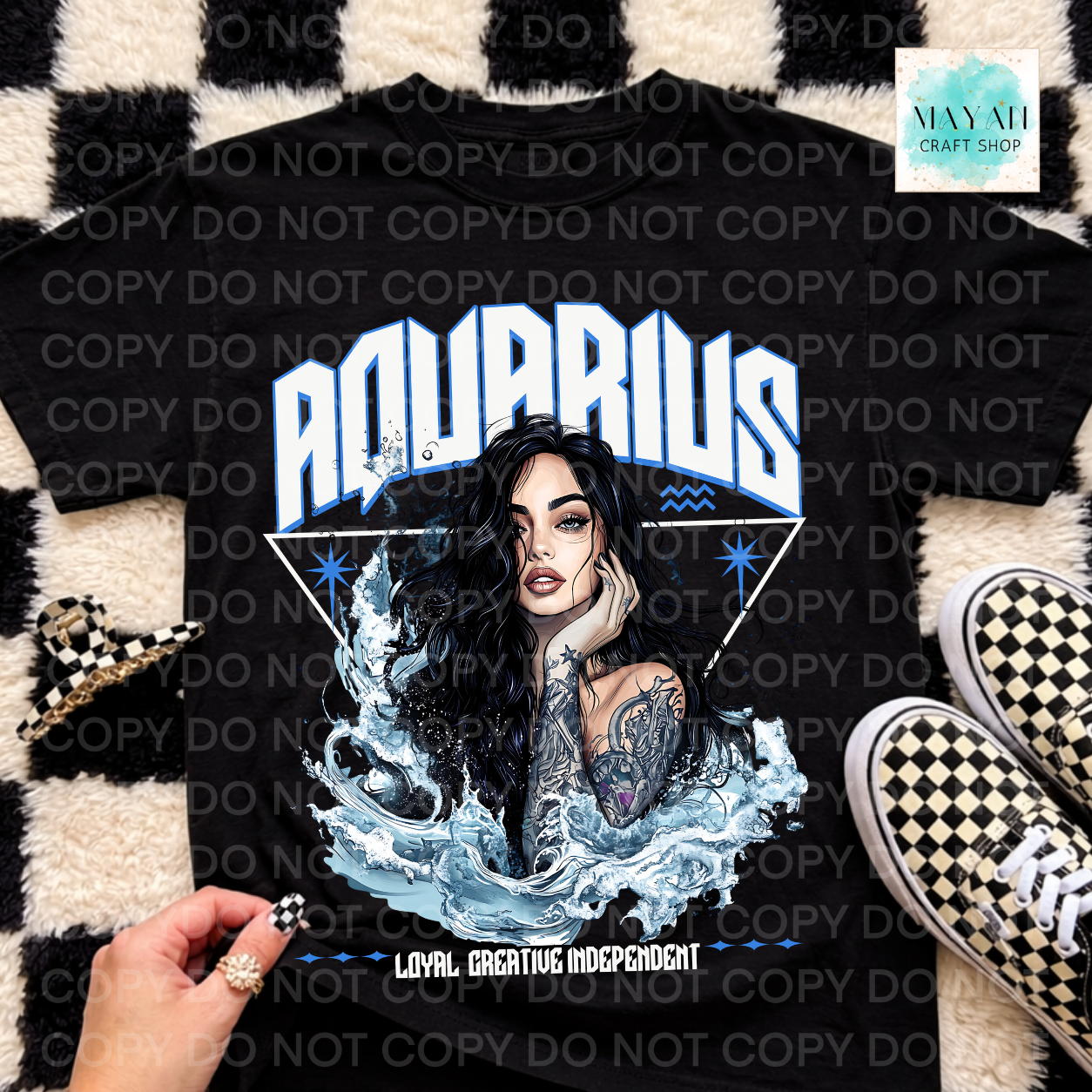 A black graphic t-shirt featuring an Aquarius-themed illustration with a woman's portrait, water, and the text 'AQUARIUS' and 'LOYAL CREATIVE INDEPENDENT'. The shirt is displayed in a flat-lay arrangement on a black and white checkered rug, next to a hand, a small checkered accessory, and black-and-white checkered sneakers.