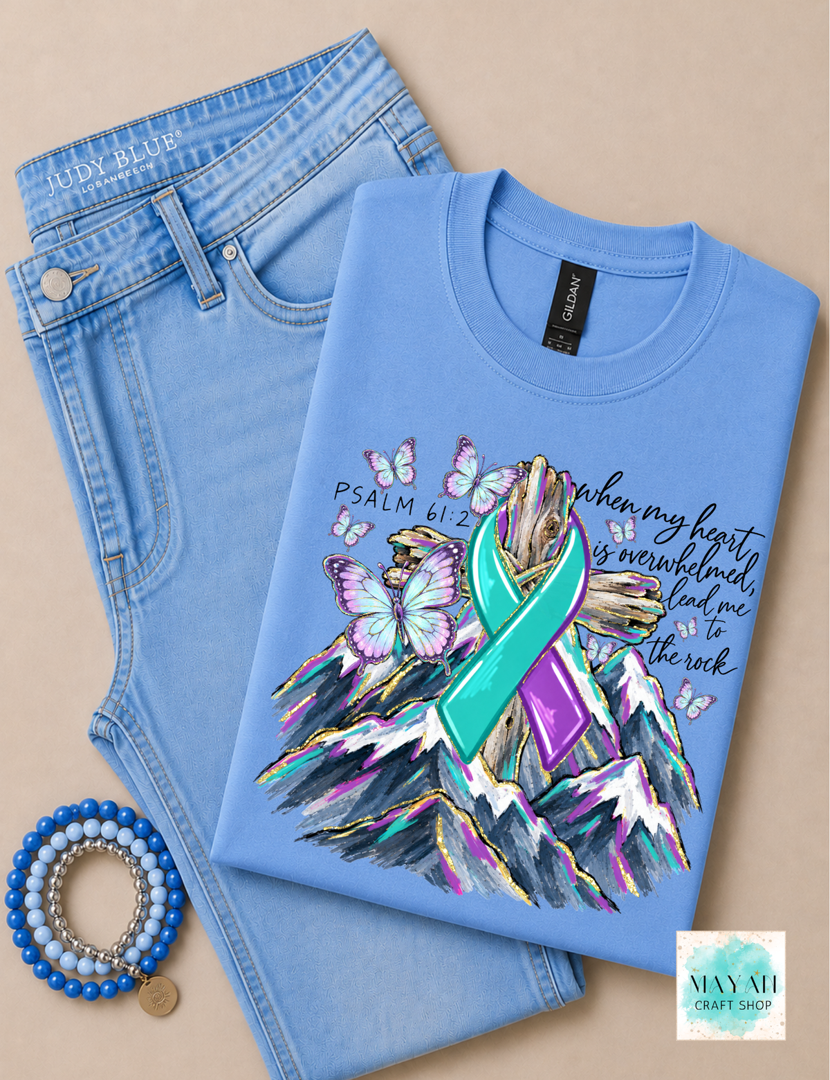 A folded light blue graphic T-shirt displayed on top of light blue jeans, with a beaded bracelet nearby on a beige background. The shirt features a colorful graphic of a teal and purple awareness ribbon over a mountain and butterfly design, with a text and verse Psalm 61:2.