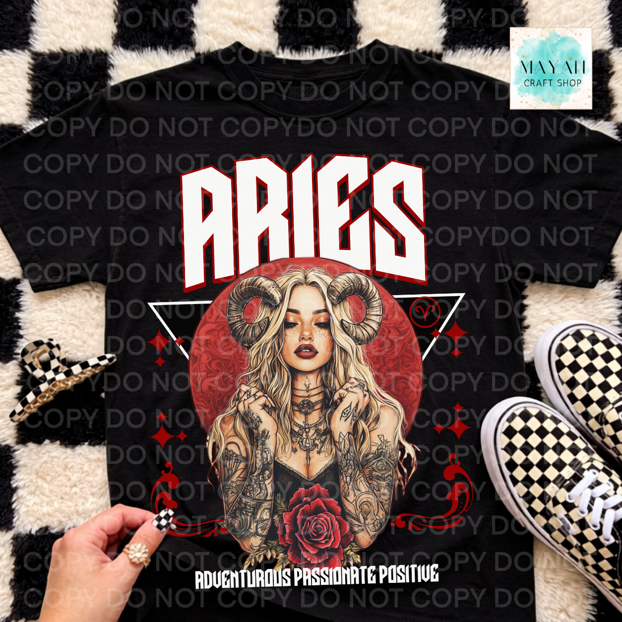 A black graphic t-shirt featuring a red circular design with a blonde woman with ram horns, the word 'ARIES' in bold white letters, and a red rose. The shirt is laid flat on a black and white checkered rug with a hand holding a checkered nail clip, and shoes.