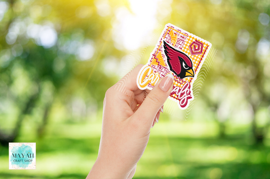 A hand holds a colorful football team logo themed sticker against a bright, blurred green park background. The sticker features a football logo in a retro font.