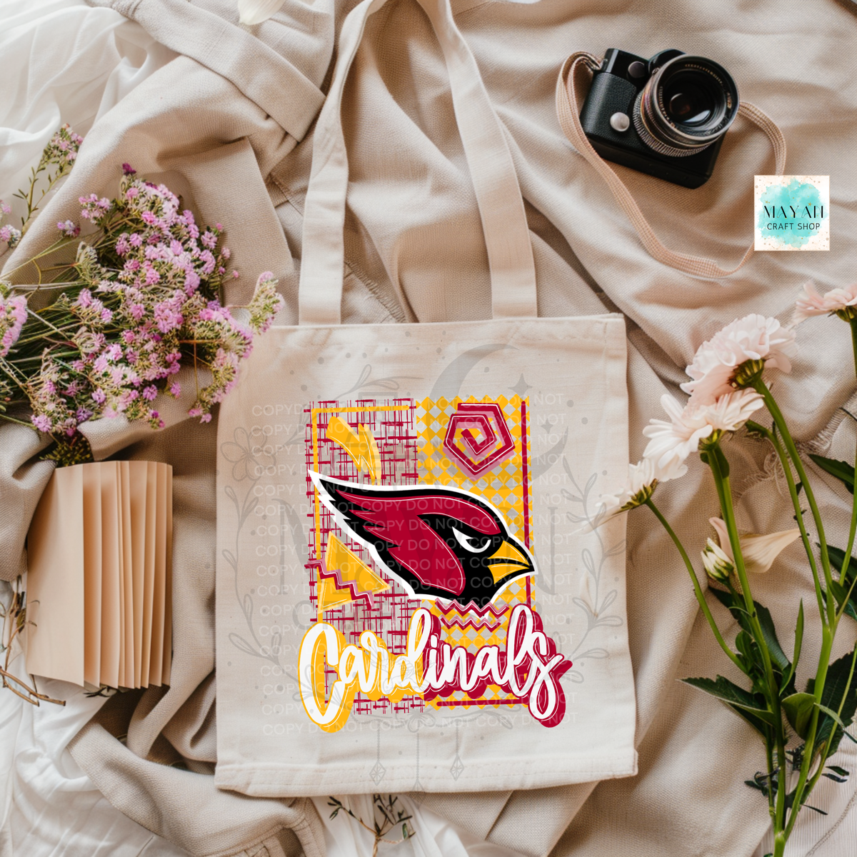 A cream canvas tote bag with a colorful football team graphic print, styled in a flat-lay arrangement with flowers, a camera, and a notebook on draped beige fabric.