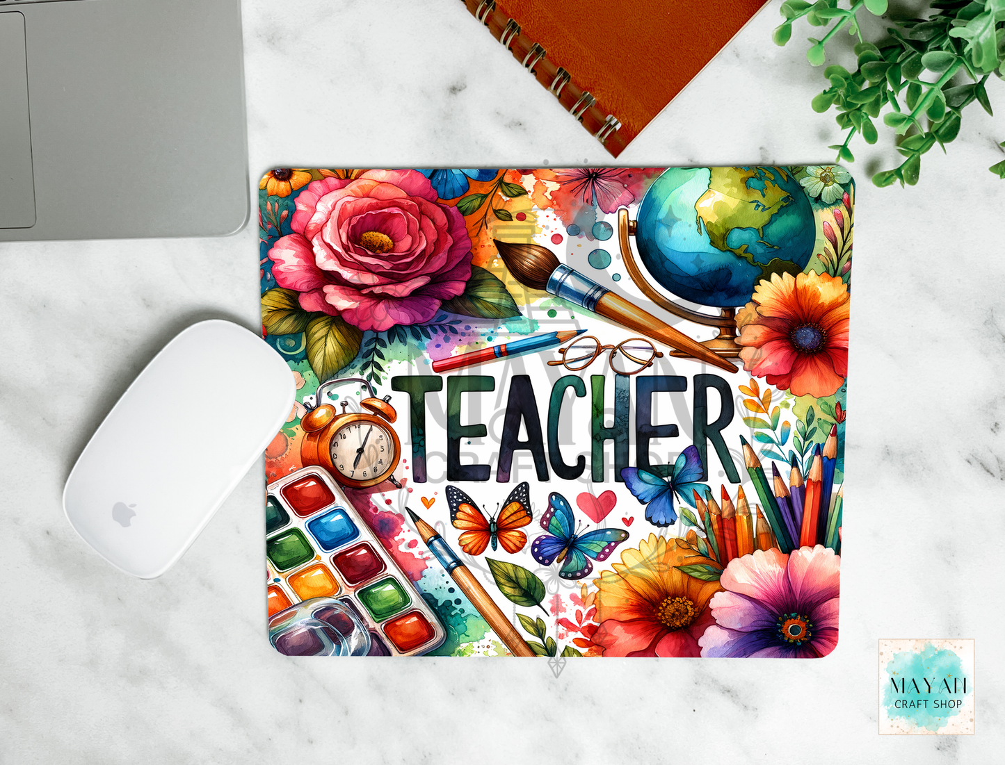 A colorful mousepad with a watercolor-style 'TEACHER' design, shown in a styled flat-lay on a white marble desk with a laptop, mouse, notebook, and plant nearby.