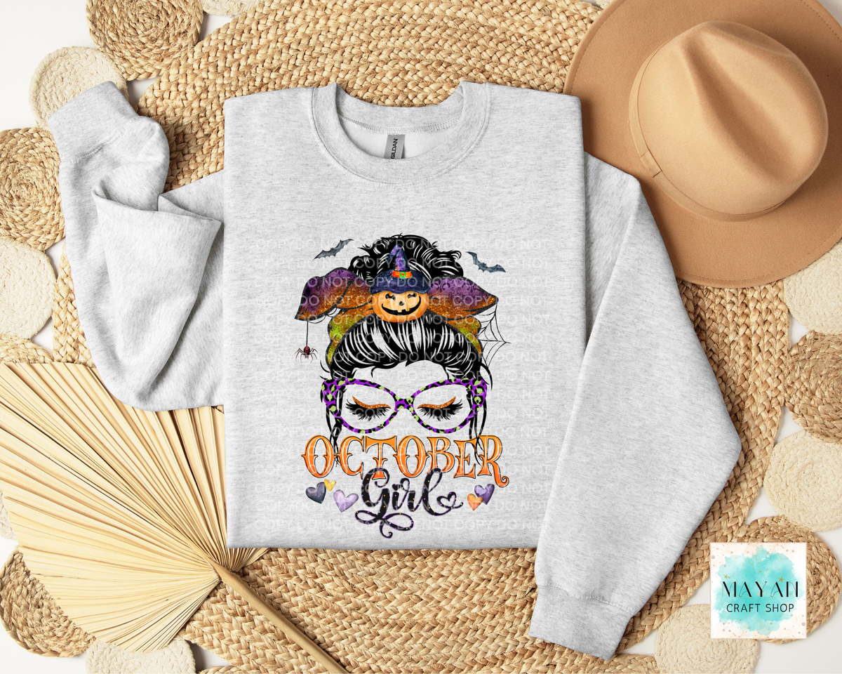 A light heather gray sweatshirt laid flat on a woven straw mat, decorated with a colorful graphic of a witch hat, pumpkin face, spiderwebs, and the words 'October Girl' in orange and purple. A tan hat, and folded napkin.