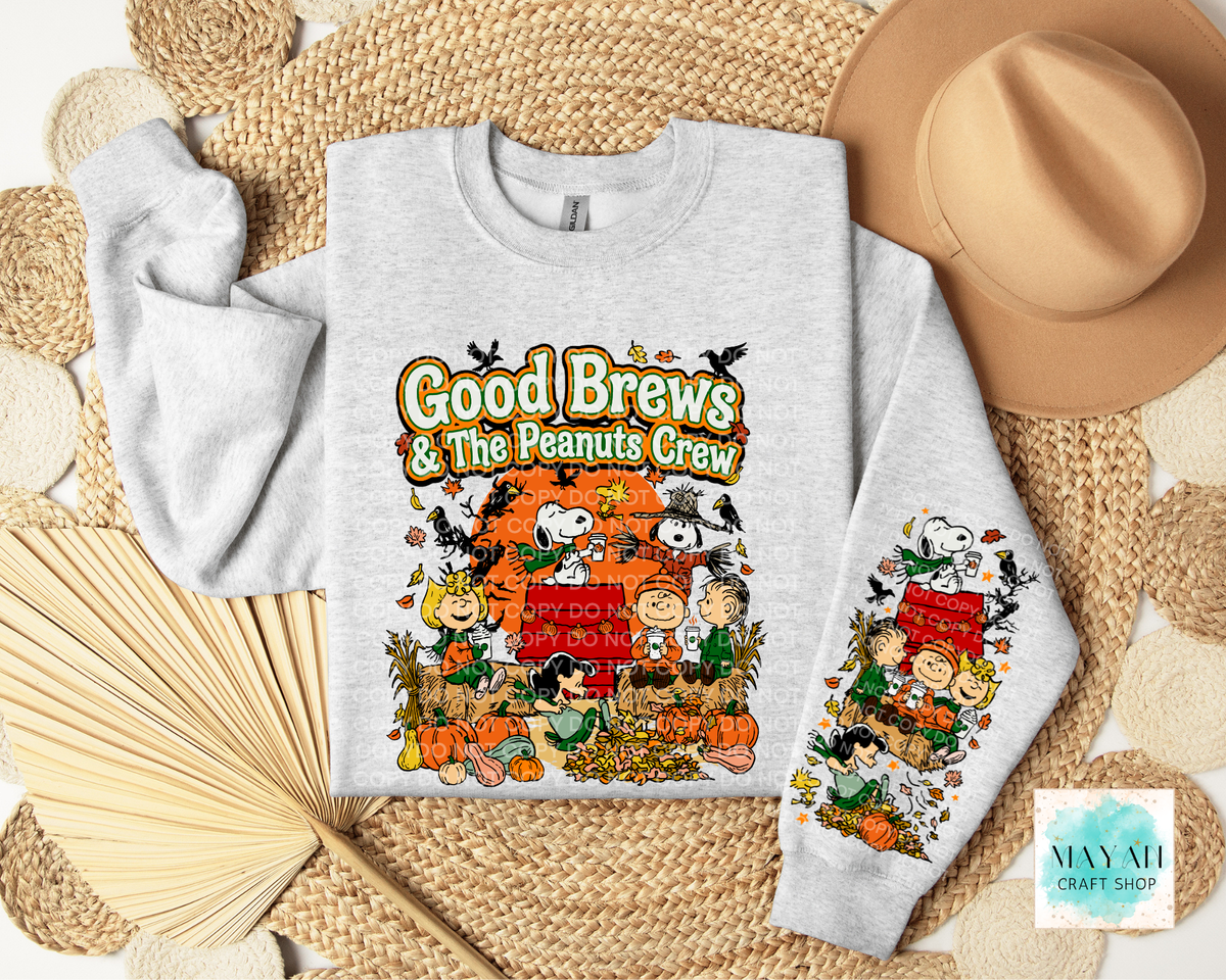 A light heather gray long-sleeve sweatshirt laid flat on a woven mat, featuring a large colorful Fall graphic on the front with the text 'Good Brews' and a cartoon scene. A matching sleeve print repeats the design.