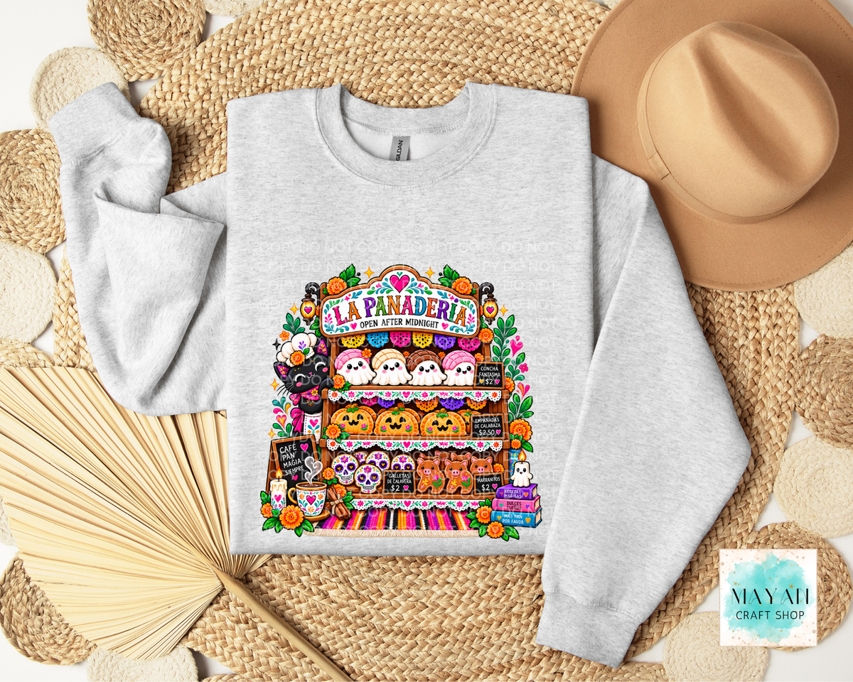 A light heather gray crewneck sweatshirt laid flat on a woven straw mat, featuring a colorful graphic of a haunted haunted house market stall with cartoon characters and pumpkins. A tan hat, folded beige towel, and wooden fan are styled around it.