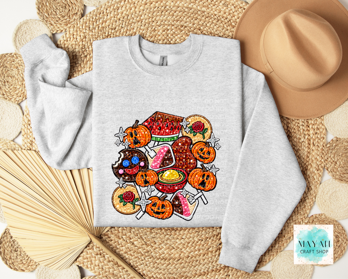 A light gray crewneck sweatshirt laid flat on a woven straw mat, featuring a colorful Halloween-themed graphic print with pumpkins, watermelon, donuts, cupcakes, and drinks. A tan hat, folded paper fans.