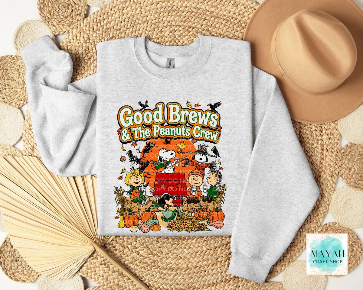 A light heather gray crewneck sweatshirt with a colorful 'Good Brews' graphic print, styled in a flat lay on a woven mat with a tan hat, paper fan.