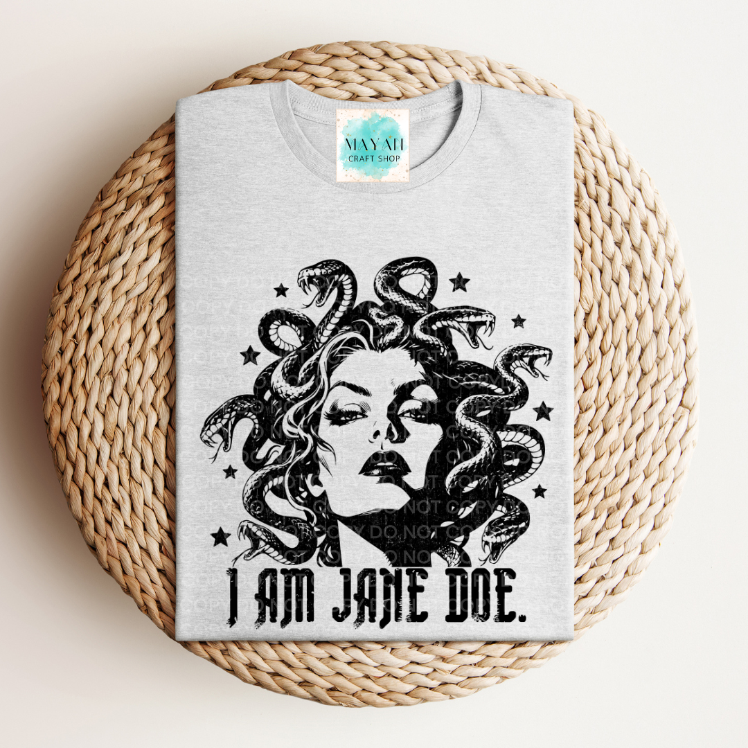 A light gray t-shirt featuring a black graphic of Medusa with the text 'I AM JANE DOE.' underneath, displayed flat on a round woven straw mat.