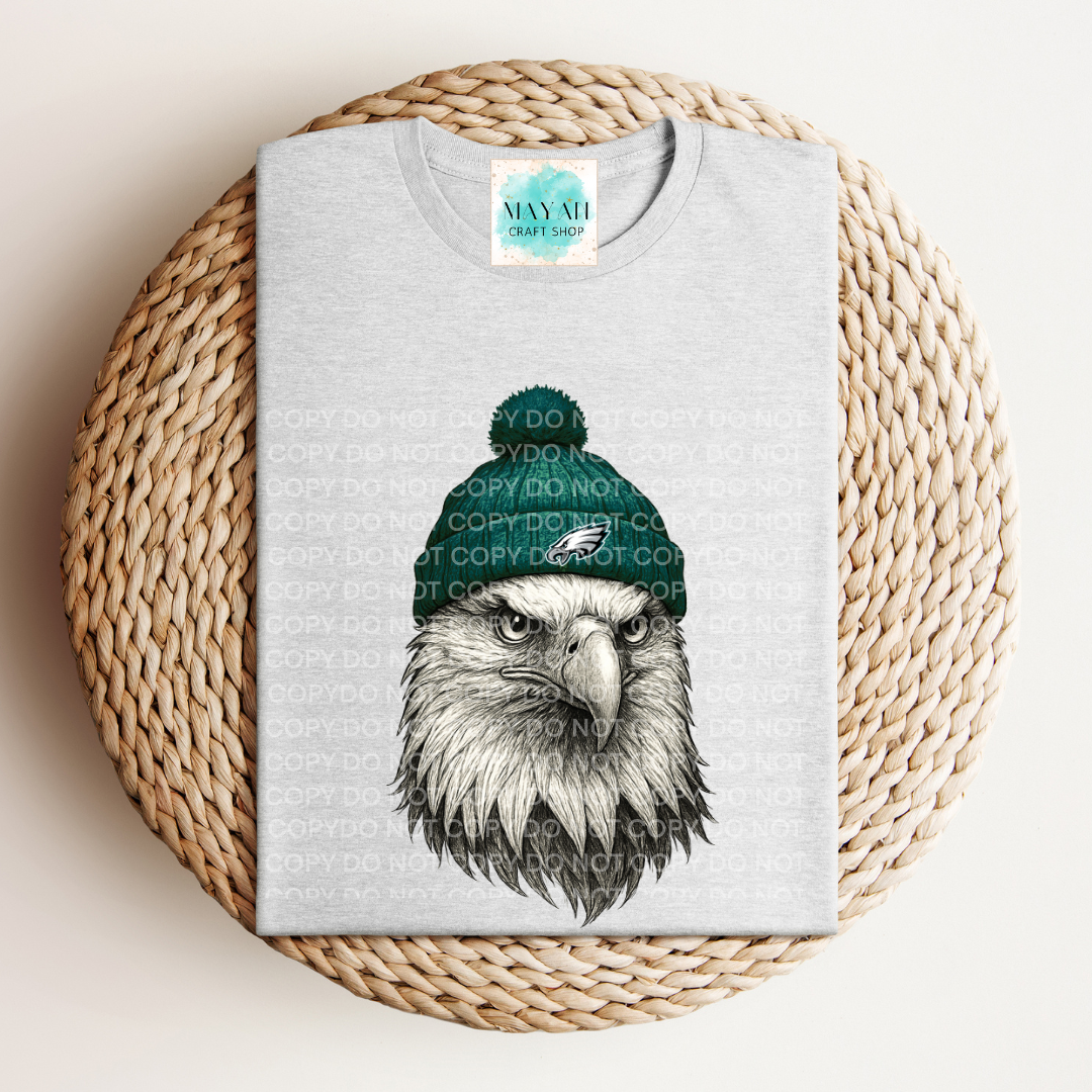 Gray t-shirt with an eagle wearing a green beanie on a woven mat