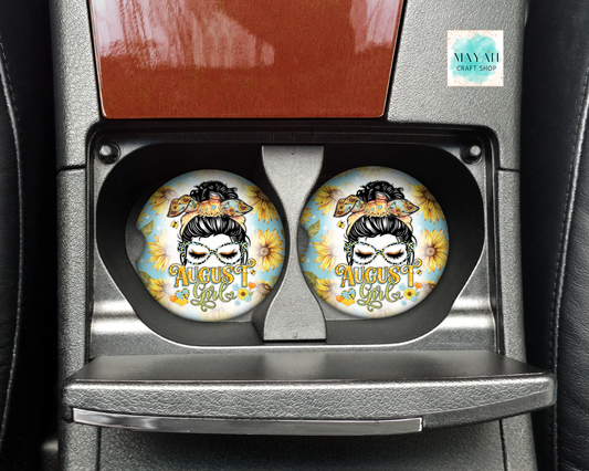 Two round car cup holders with a detailed graphic of a woman's face surrounded by sunflowers and the text 'August 1st' in gold, installed in a car's center console.