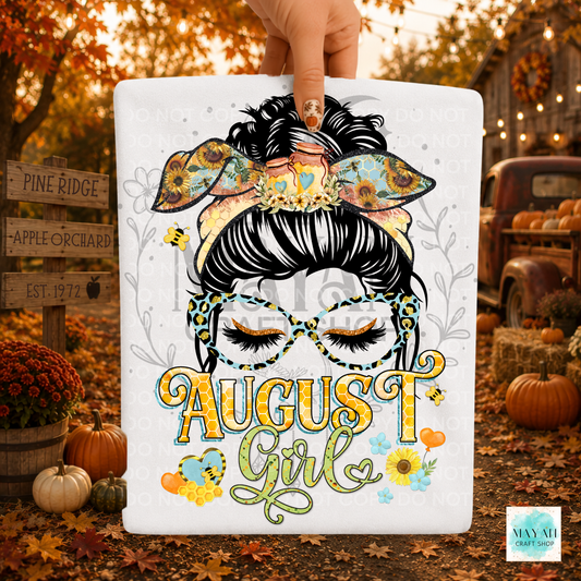 A hand holds a white rectangular sign featuring a stylized illustrated woman with black hair, patterned bow and glasses, and the words “August Girl” in autumn colors. The sign is photographed outdoors in a fall yard with pumpkins, leaves, and rustic decor in the background.