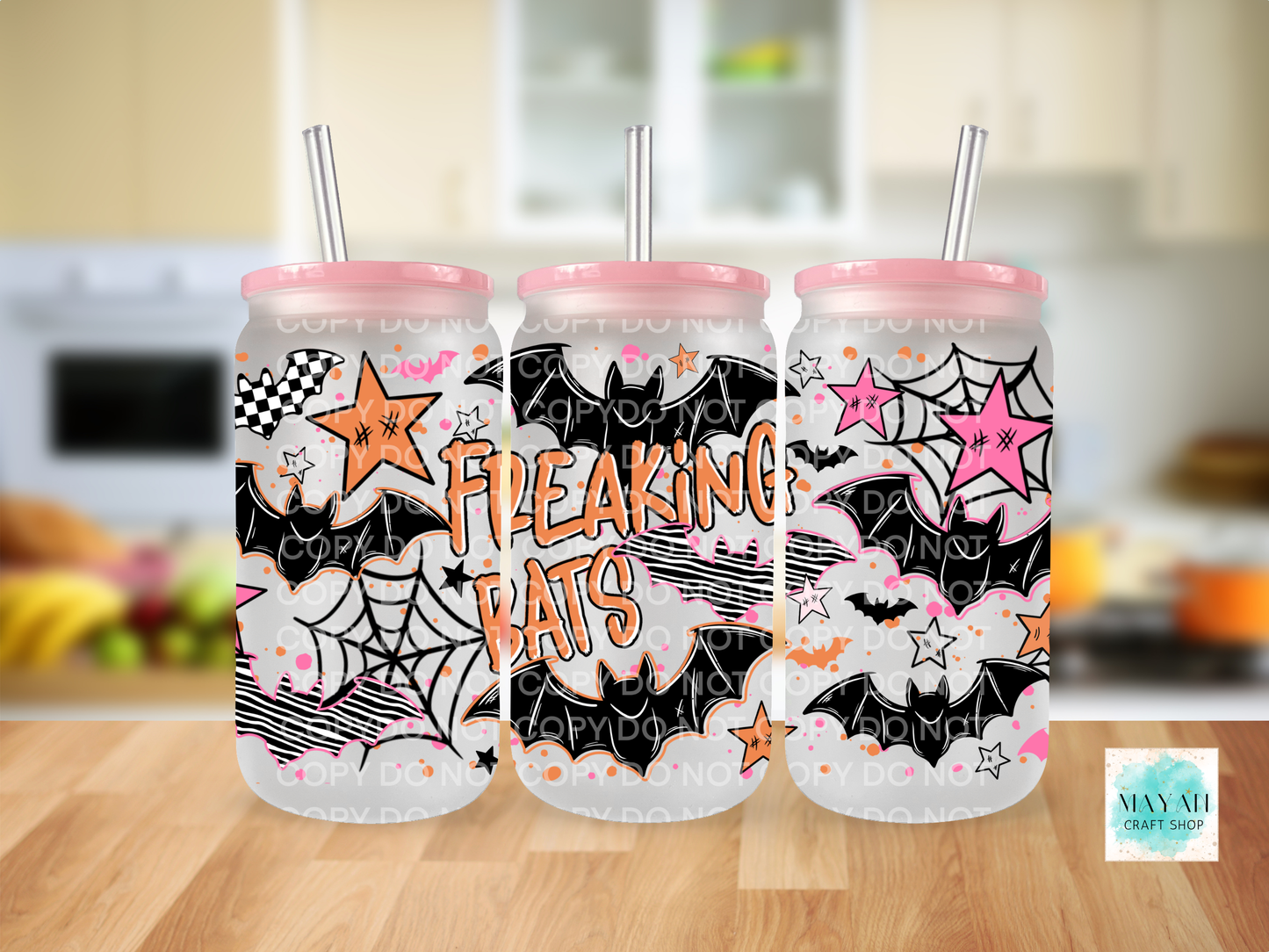Three frosted plastic tumblers with pink lids and metal straws, decorated with Halloween-themed graphics including bats, spiders, stars, and the words 'FREAKING BATS' in orange, shown on a wooden table with a blurred kitchen background.