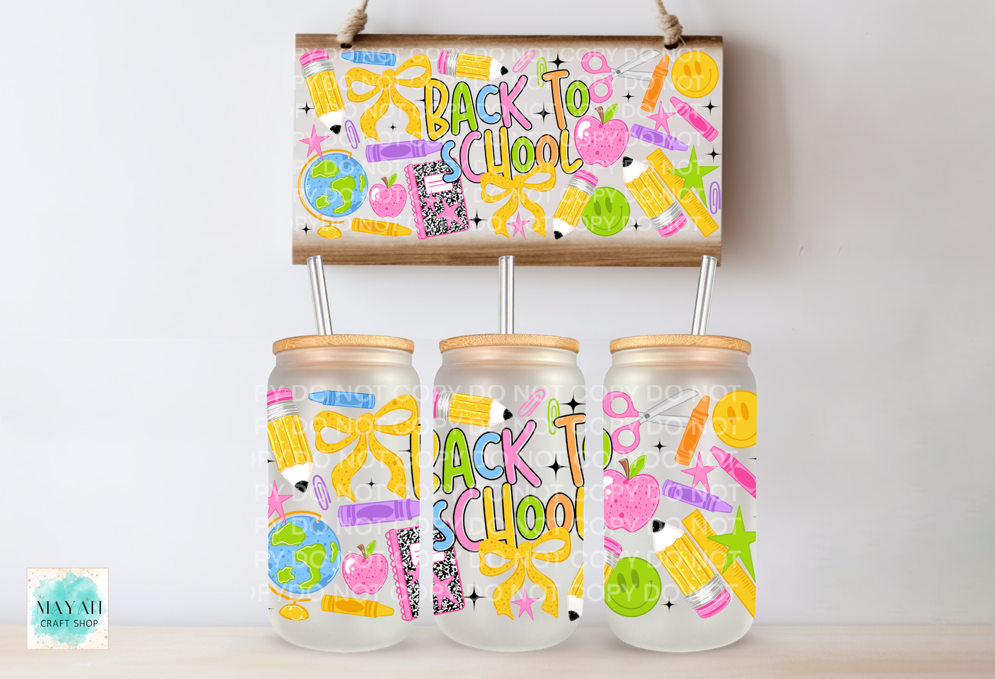 Three clear glass can with bamboo lids and metal straws are displayed below a matching wooden sign decorated with colorful school supplies and the words 'Back to School.' The jars and sign feature playful multicolor graphics including bows, pencils, stars, smiley faces, and small illustrations.