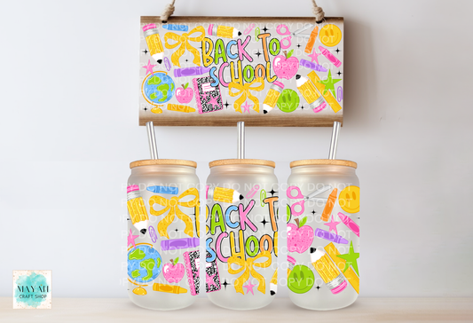 Three clear glass can with bamboo lids and metal straws are displayed below a matching wooden sign decorated with colorful school supplies and the words 'Back to School.' The jars and sign feature playful multicolor graphics including bows, pencils, stars, smiley faces, and small illustrations.
