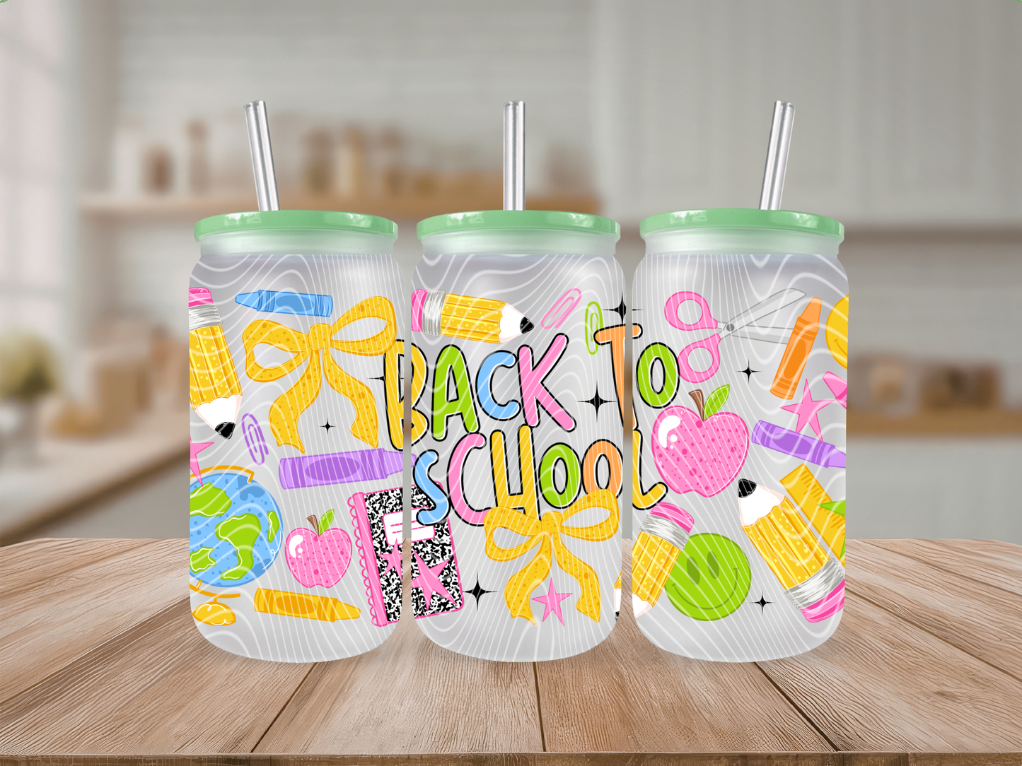Three frosted glass tumblers with mint green lids and clear straws, decorated with colorful 'Back to School' illustrations including bows, pencils, apples, and books, displayed on a wooden table with a blurred kitchen background.