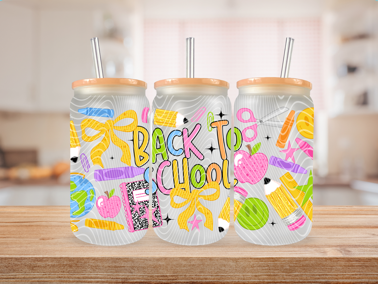 Three frosted glass cans with orange lids and metal straws, decorated with colorful school-themed illustrations and the words “BACK TO SCHOOL,” displayed on a wooden surface with a blurred kitchen background.