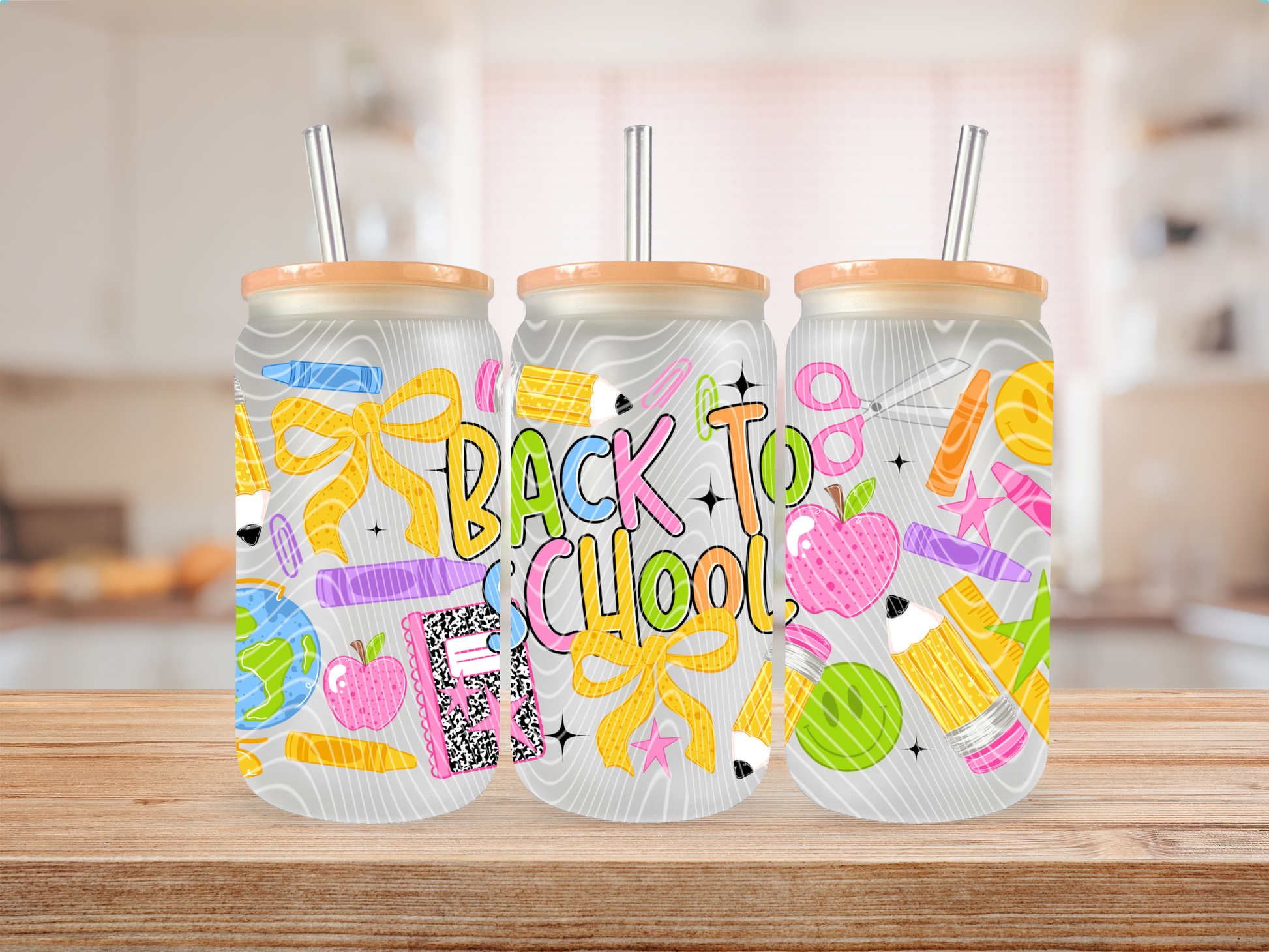 Three frosted glass cans with orange lids and metal straws, decorated with colorful school-themed illustrations and the words “BACK TO SCHOOL,” displayed on a wooden surface with a blurred kitchen background.