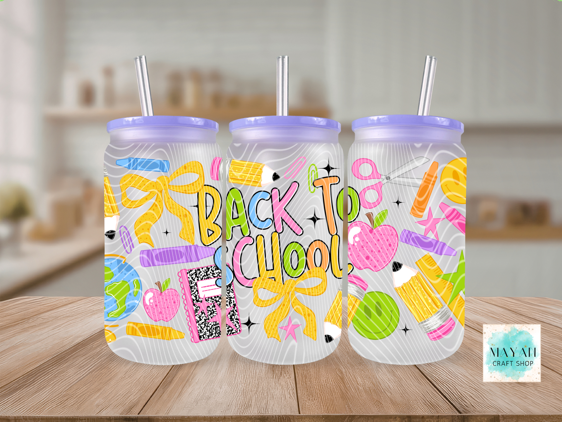 Three clear plastic tumblers with purple lids and metal straws, decorated with colorful school-themed graphics and the words 'BACK TO SCHOOL' in pastel letters, displayed on a wooden table with a blurred kitchen background.