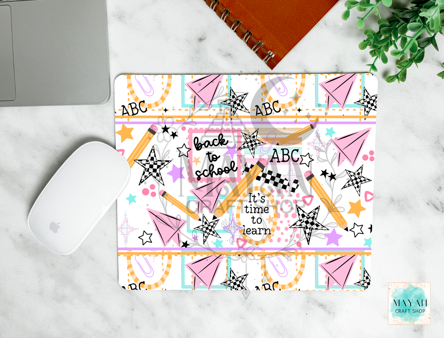 A mouse pad with a colorful 'Back to School' theme, featuring pencils, stars, paper airplanes, and the text 'It's time to learn' and 'ABC', shown on a white marble desk with a mouse, laptop corner, notebook, and plant.