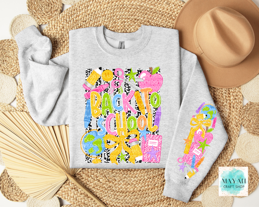A light heather gray sweatshirt laid flat on a woven straw mat, decorated with a colorful 'Back to School' graphic on the front and a matching graphic on the sleeve. A tan hat, folded fan, and woven mat surround it.