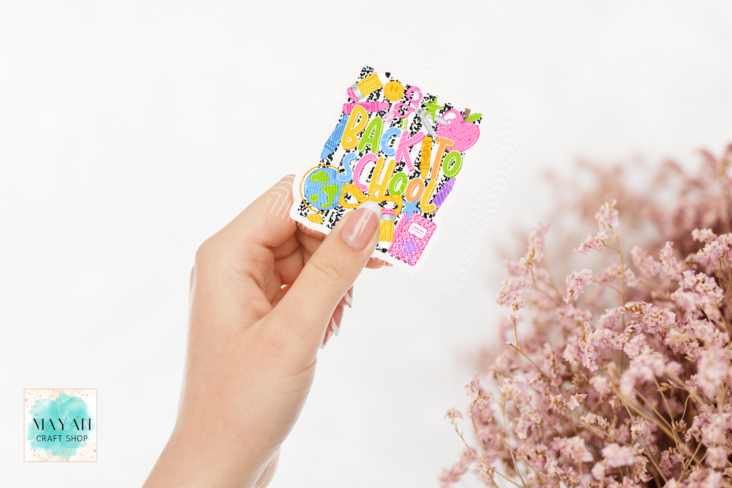 A hand holds a small square sticker sheet featuring colorful cartoon back to school themed. The background is a clean white with soft pink dried flowers on the right and a 'May Ali Craft Supply' logo watermark in the bottom-left corner.