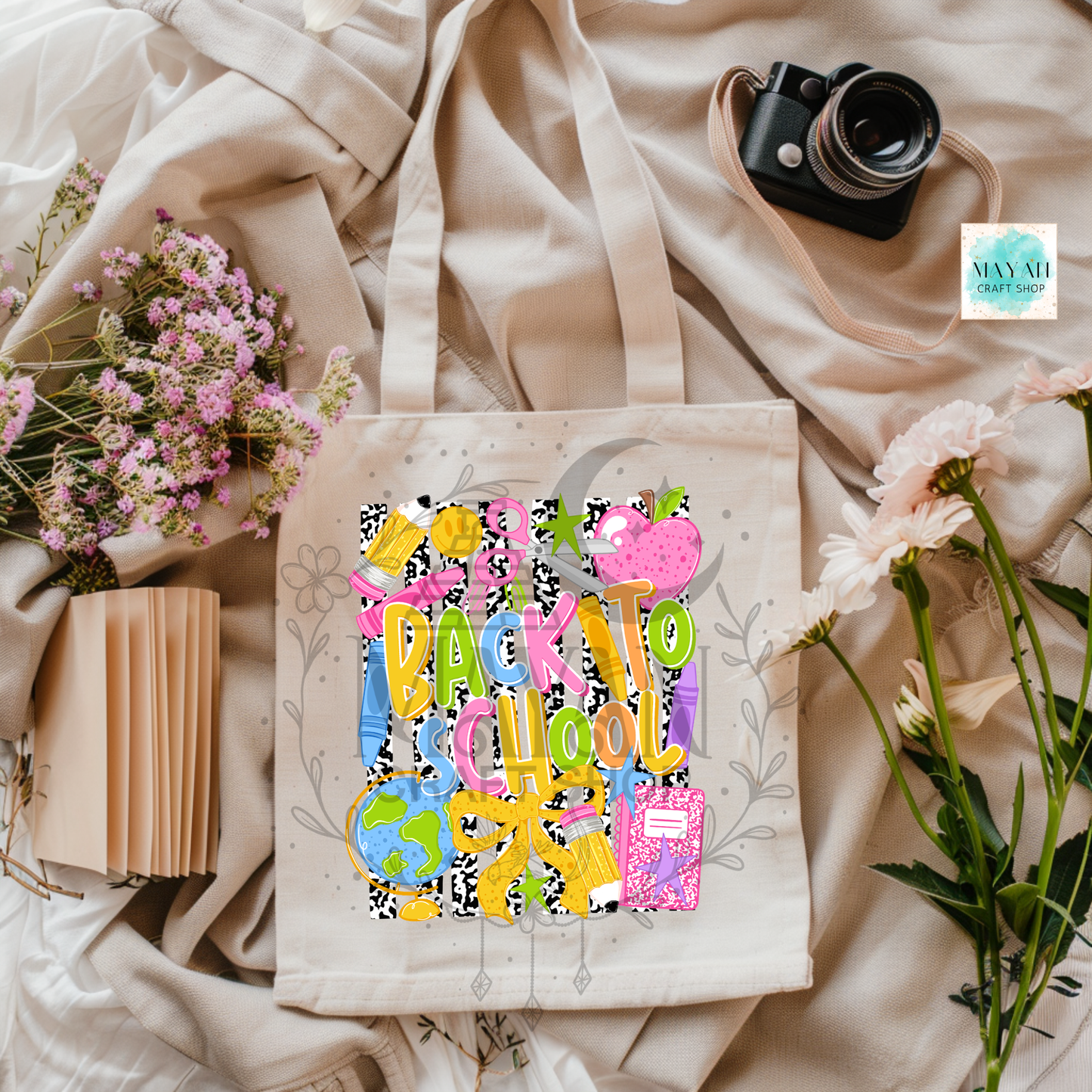 A cream canvas tote bag with a colorful 'Back to School' graphic print, styled in a flat-lay arrangement with a vintage camera, pink apples, a book, and draped beige fabric.