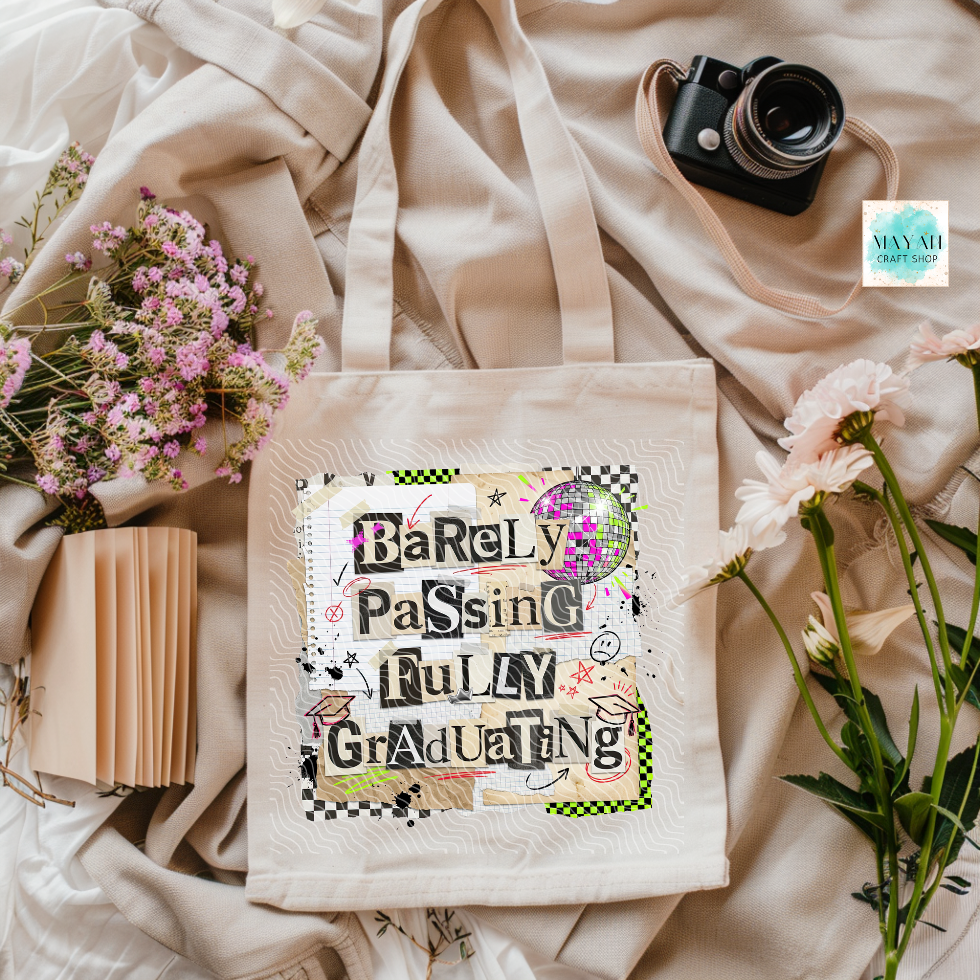 Tote bag with 'Barley Passing Fully Graduating' text on a soft fabric background with flowers and a camera.