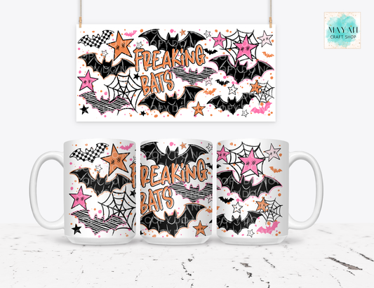 A white ceramic mug shown from three angles with a matching hanging banner above it, both printed with a Halloween-style design featuring black bats, pink and orange stars, spiderwebs, and the words “FREAKING BATS.”.