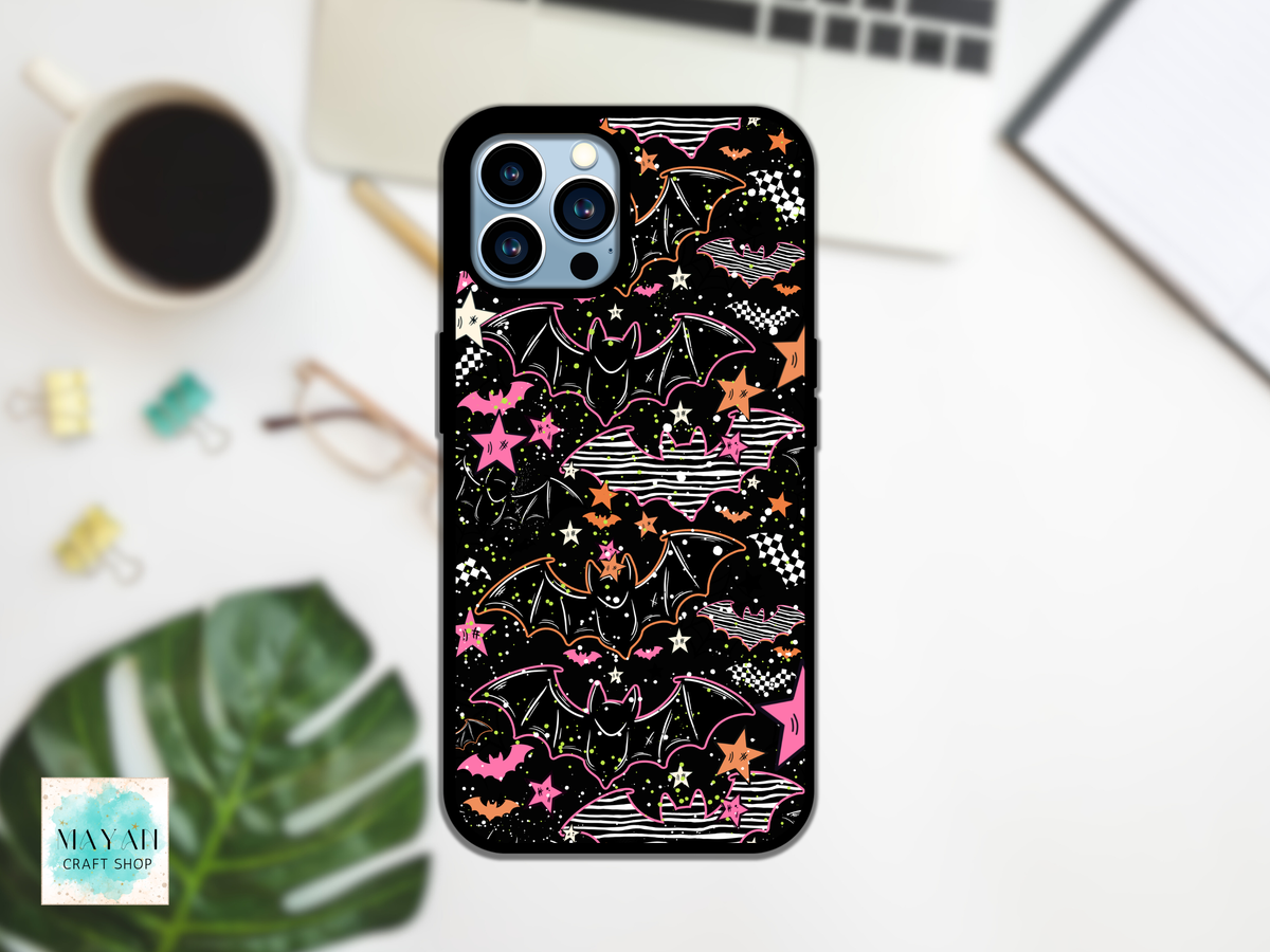 A phone case with a black background and a colorful Halloween-themed pattern featuring bats, stars, and clouds in pink, orange, and white, shown on a phone in a styled desk flat-lay setting.