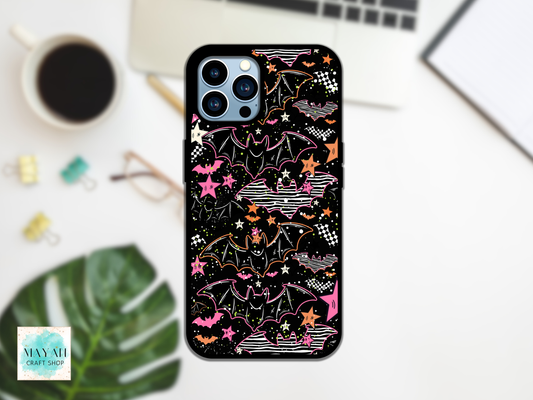 A phone case with a black background and a colorful Halloween-themed pattern featuring bats, stars, and clouds in pink, orange, and white, shown on a phone in a styled desk flat-lay setting.