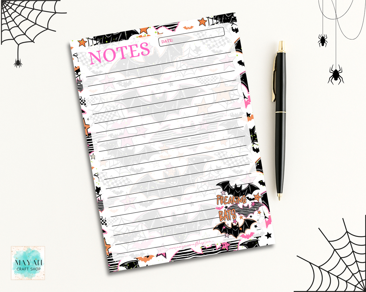 A Halloween-themed notepad with lined writing space, pink 'NOTES' text, bat and spider graphics, and a black pen placed beside it on a light background with spiderweb decorations.