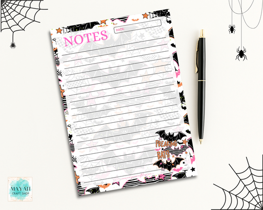 A Halloween-themed notepad with lined writing space, pink 'NOTES' text, bat and spider graphics, and a black pen placed beside it on a light background with spiderweb decorations.