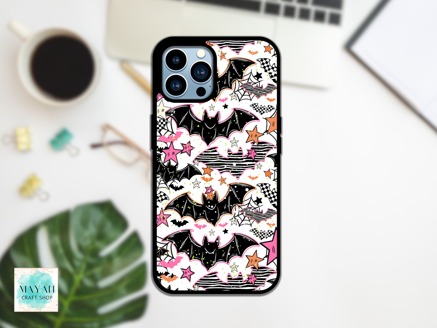 A phone case featuring a Halloween-themed pattern of black bats, pink stars, and white clouds with black line art, shown on a white smartphone. The case is photographed in a styled flat-lay setting on a white desk with a coffee cup, laptop, paper clips, glasses, a green leaf, and a notebook in the background.