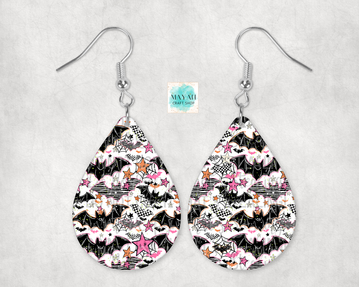 A pair of teardrop-shaped dangle earrings with a black, white, and pink bat-themed pattern, hanging from silver fishhook wires on a light gray textured background.