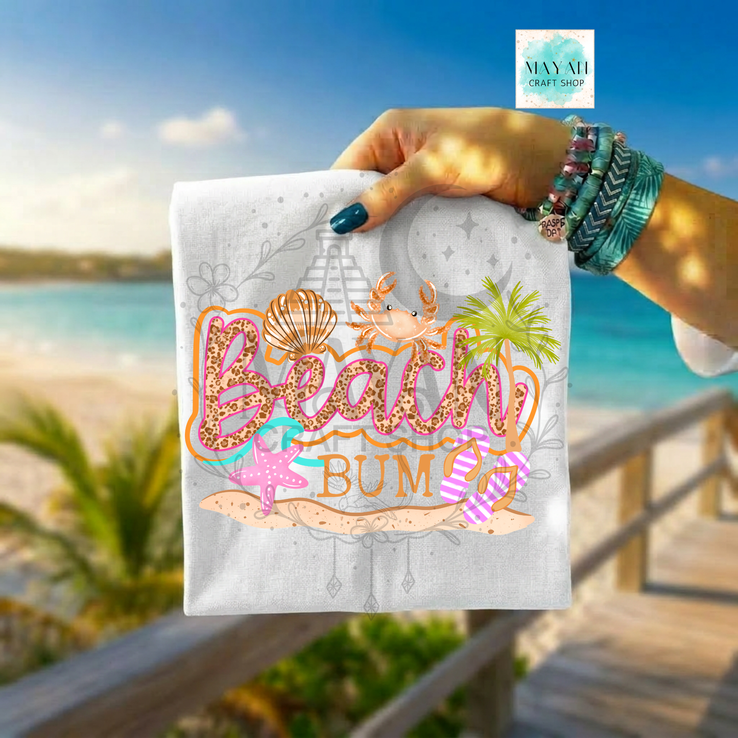 Hand holding a towel with 'Beach Bum' design against a beach backdrop