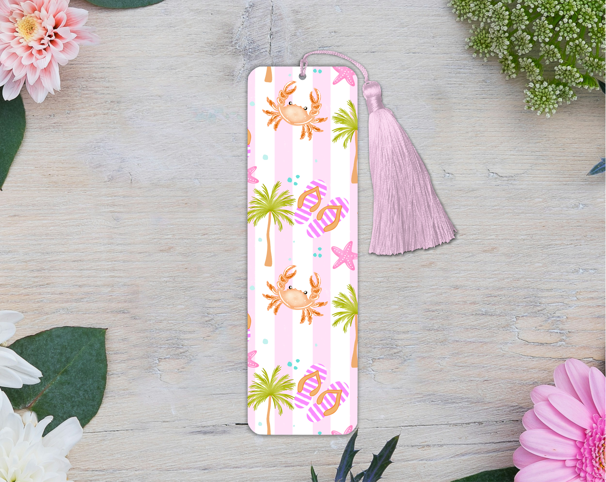 Bookmark with crab and palm tree pattern on a pink background with a tassel, placed on a wooden surface with flowers.