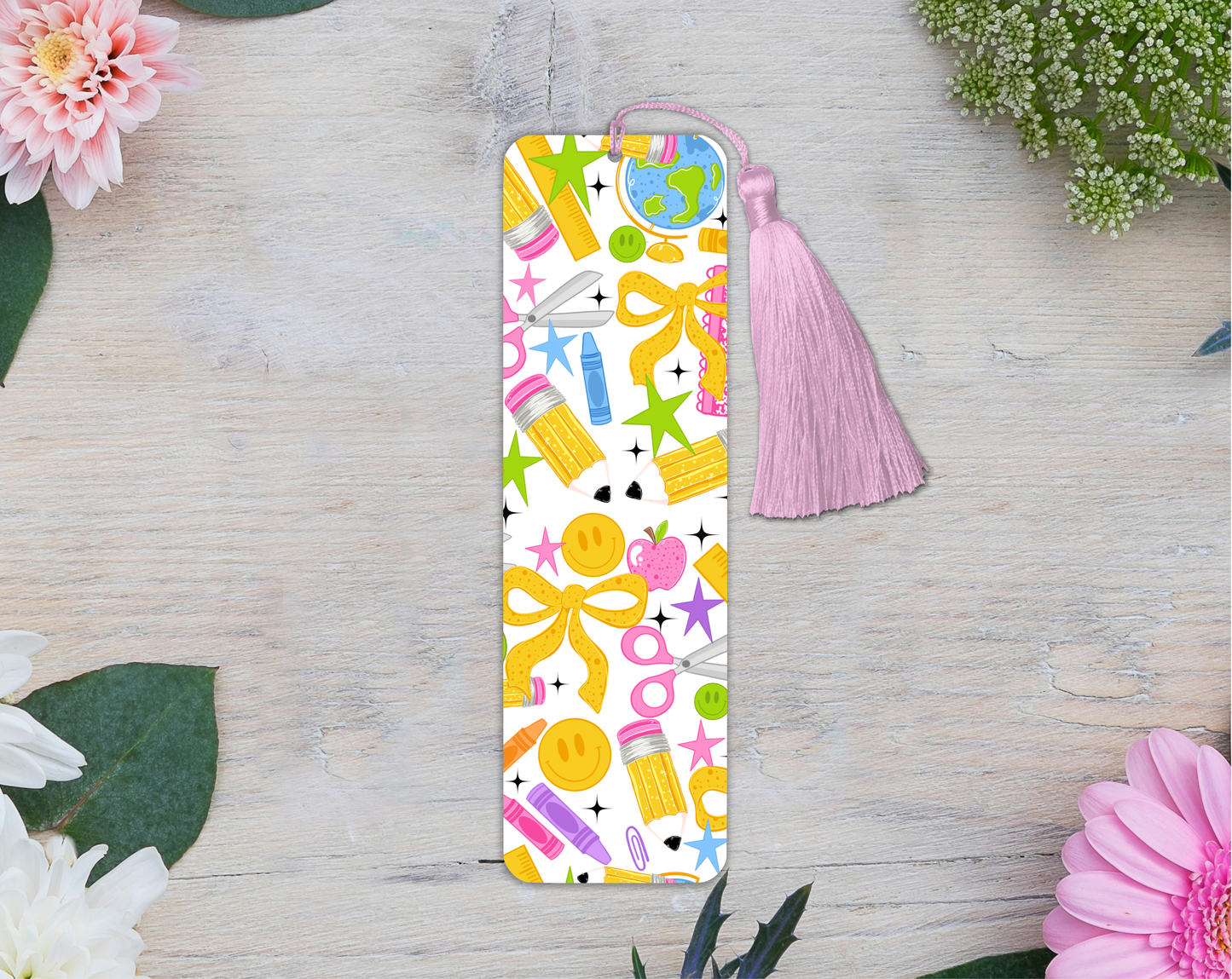 A slim rectangular bookmark with a pink tassel, featuring a colorful pattern of school supplies and bows on a white background, styled on a light wood surface with flowers.