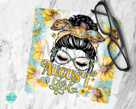 A lens cloth cleaner featuring a stylized illustration of a woman with black hair in a messy bun, wearing patterned sunglasses and large decorative glasses, with the words “August Girl” in gold script. The design includes yellow sunflowers and blue accents on a light background, shown on a white marble surface with a pair of black eyeglasses resting on top.