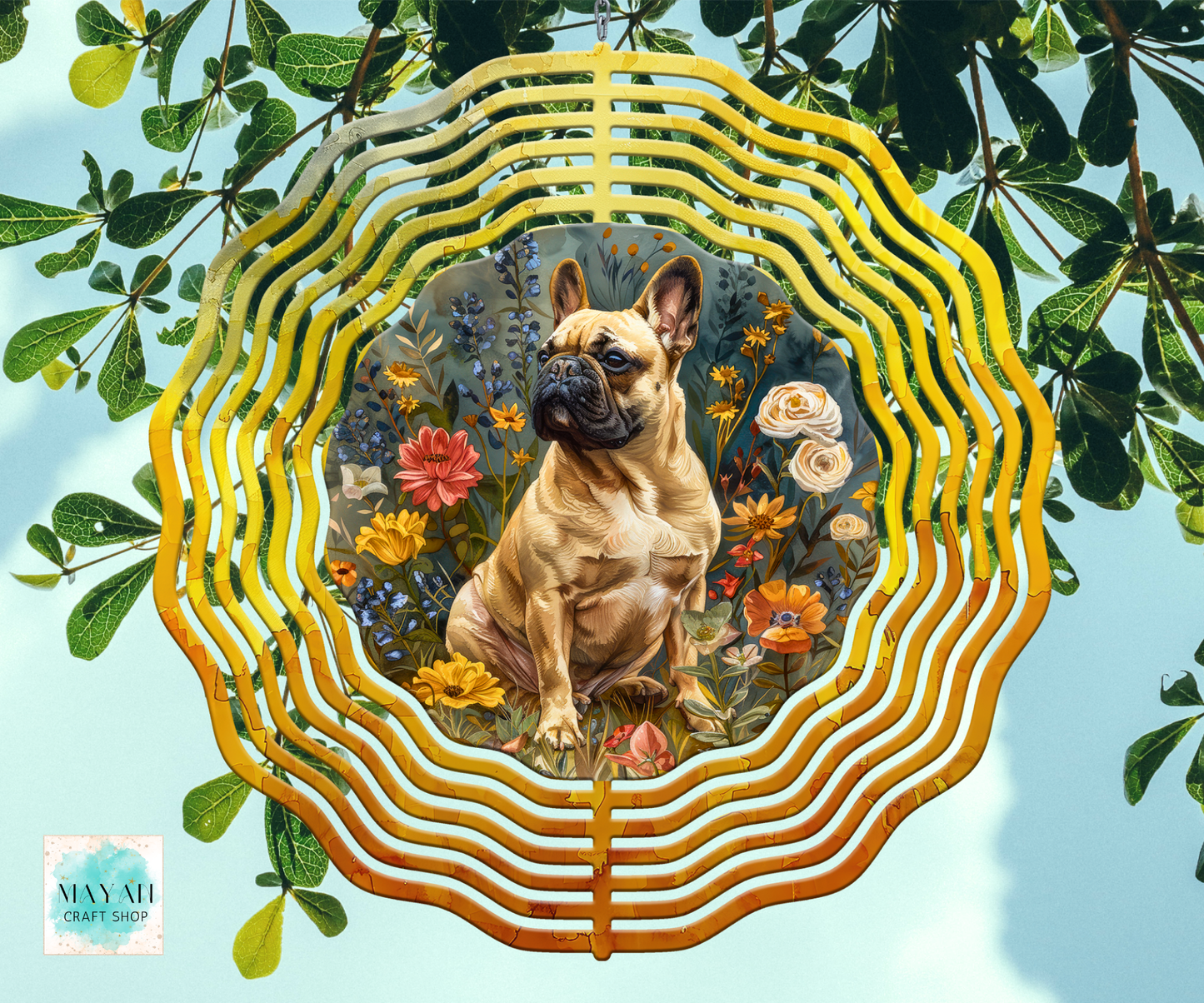 A wind spinner hanging or ornament featuring a painted French bulldog surrounded by flowers, suspended against a light blue sky background with green leaves around it. The product has a wavy, layered circular design with a gold-toned outer frame and a multicolored central illustration.
