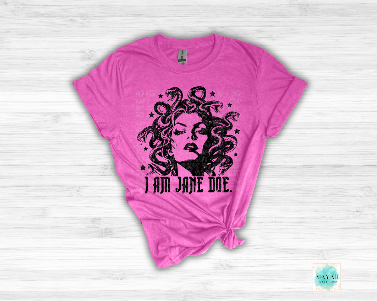 A heather berry short-sleeve t-shirt with a black graphic of Medusa's face surrounded by snakes and the text 'I AM JANE DOE.' on a light wood background.