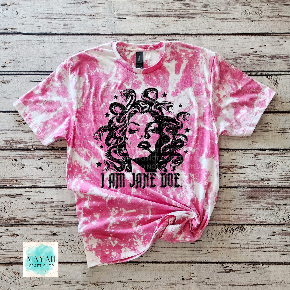 A heather berry bleached graphic t-shirt featuring a black illustration of Medusa and the text 'I AM JANE DOE.' laid flat on a rustic wooden plank background.