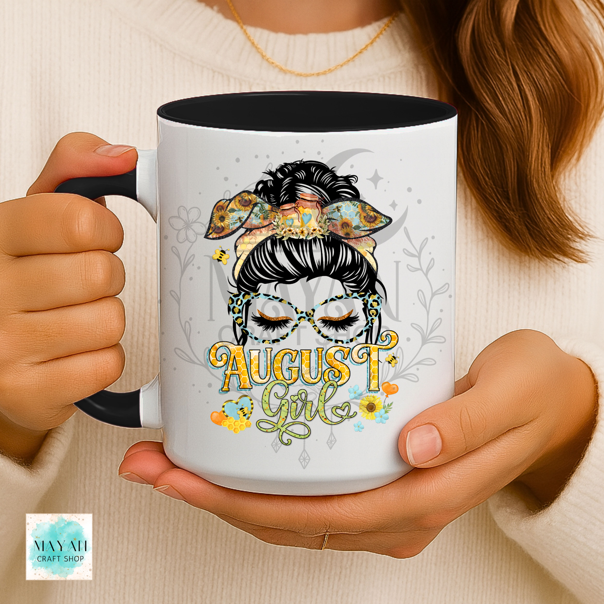 A person holds a white ceramic mug with a black interior and handle. The mug features a colorful graphic of a stylized woman with a bow and mask, with the text 'August Girl' in yellow and green.