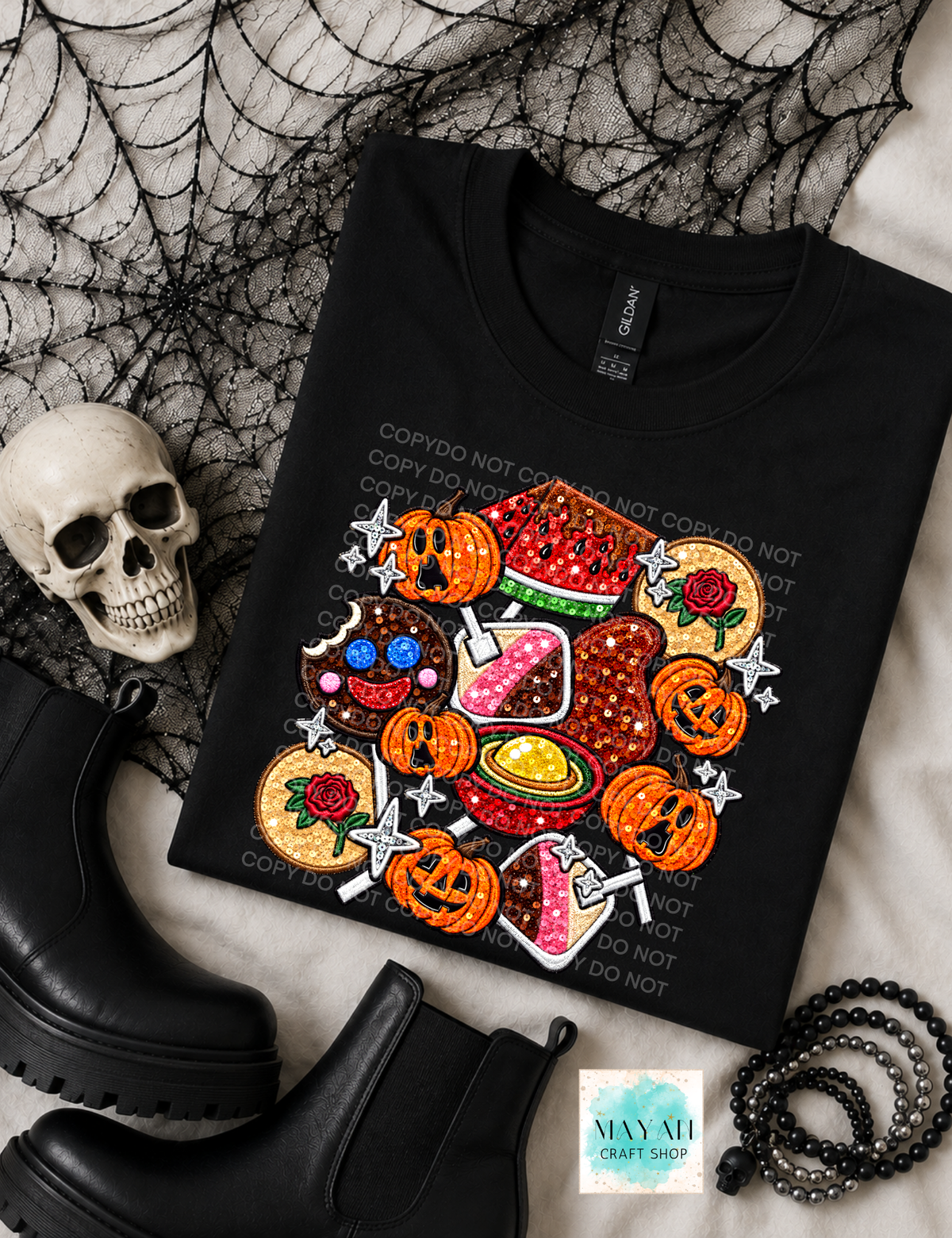 A black short-sleeve T-shirt laid flat with a colorful Halloween-themed graphic print featuring mexican candy and stars. The shirt is styled with black boots, a skull, spiderweb fabric, and a beaded bracelet on a light surface.