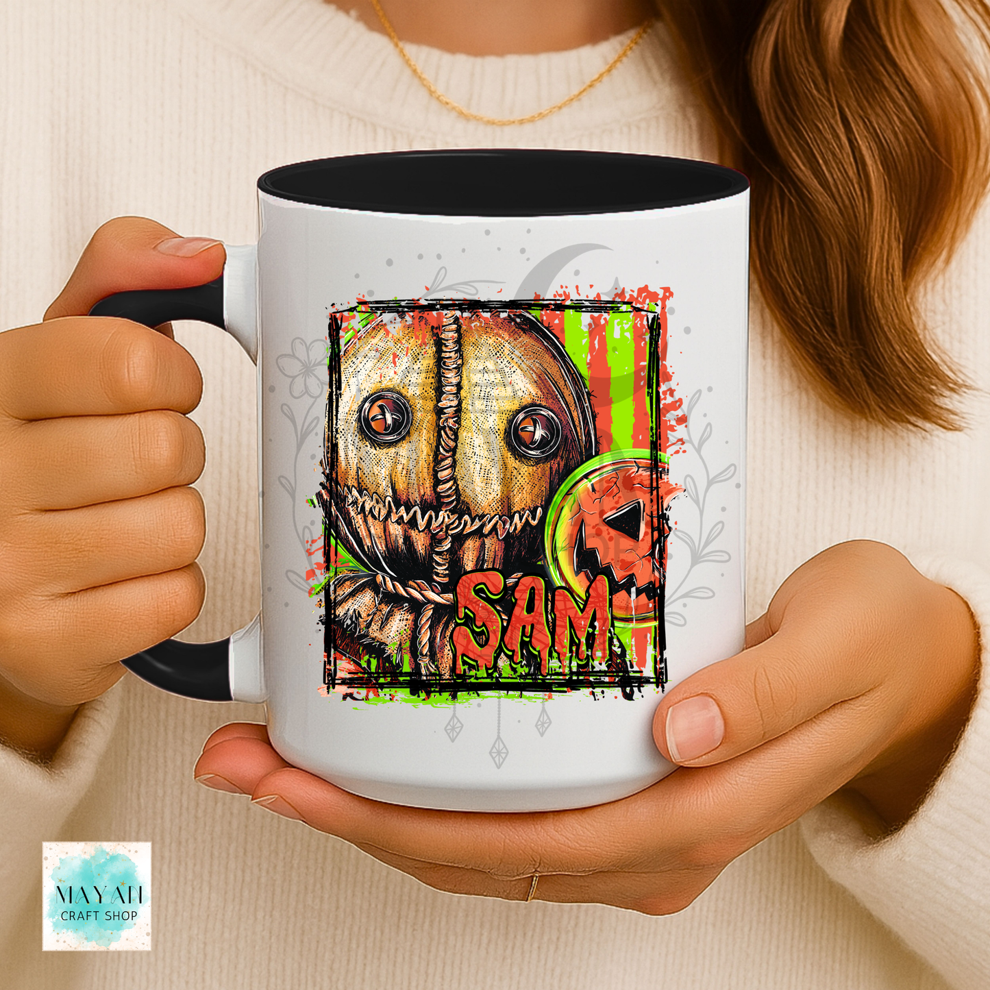 A person holds a white ceramic mug with a black handle and interior. The mug features a colorful graphic of a stitched horror character with a name in red, plus splatter and green vertical streaks.