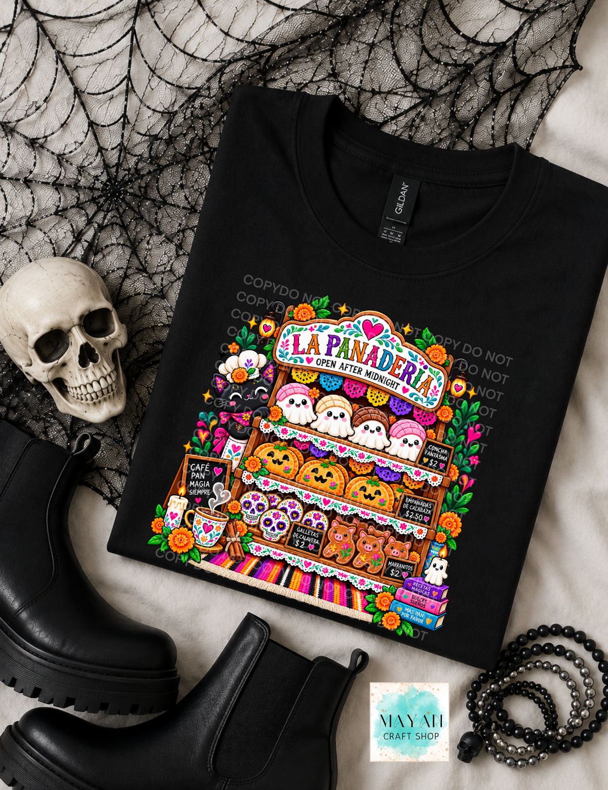 A black graphic T-shirt laid flat on a light fabric surface with Halloween-themed props, including a skull, black boots, spiderweb backdrop, and beaded bracelet. The shirt features a colorful illustrated storefront sign reading 'La Panaderia' with ghosts, pumpkins, flowers, and seasonal motifs.