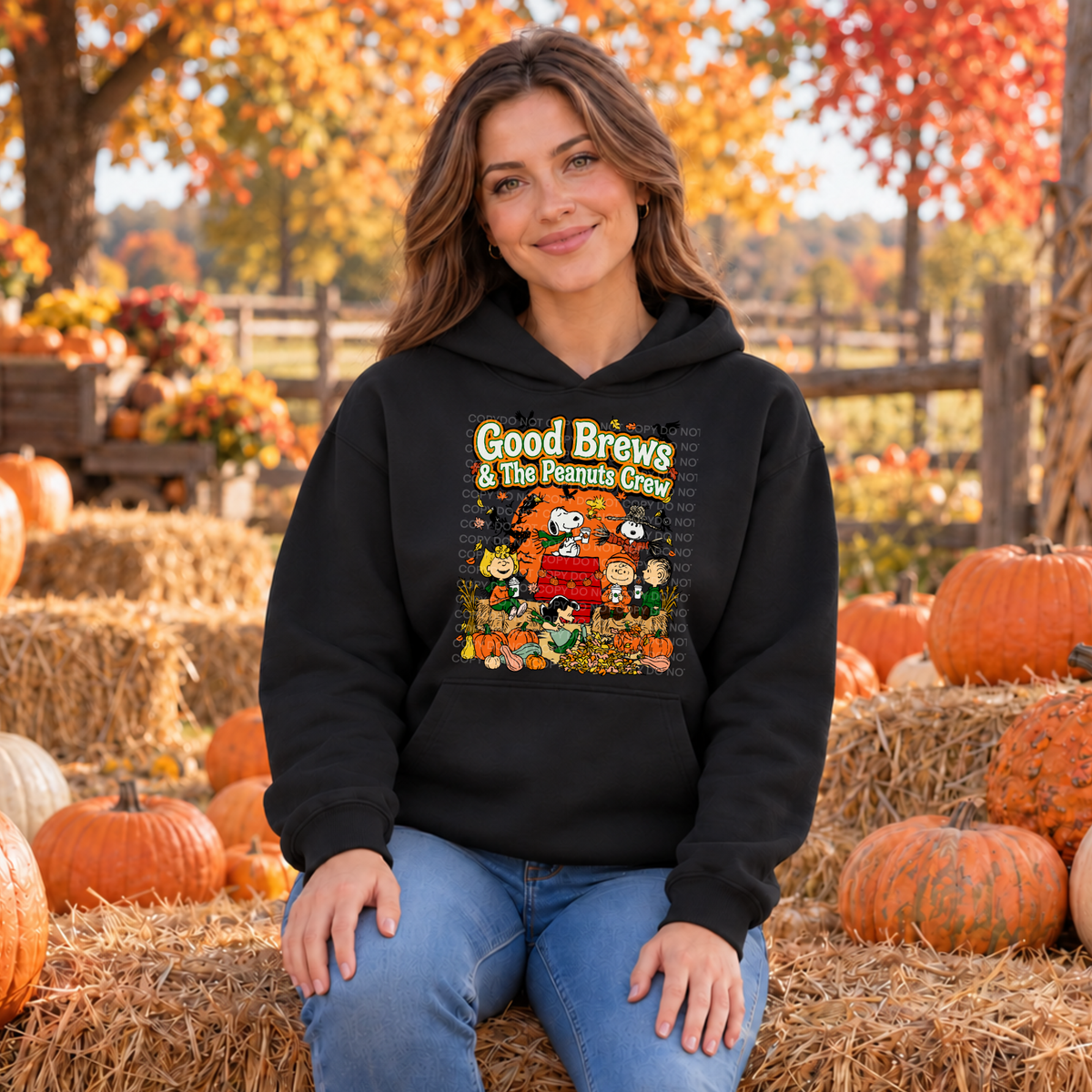A woman wearing a black hoodie with a colorful 'Good Brews' graphic print, sitting on a hay bale in an autumn pumpkin farm setting.