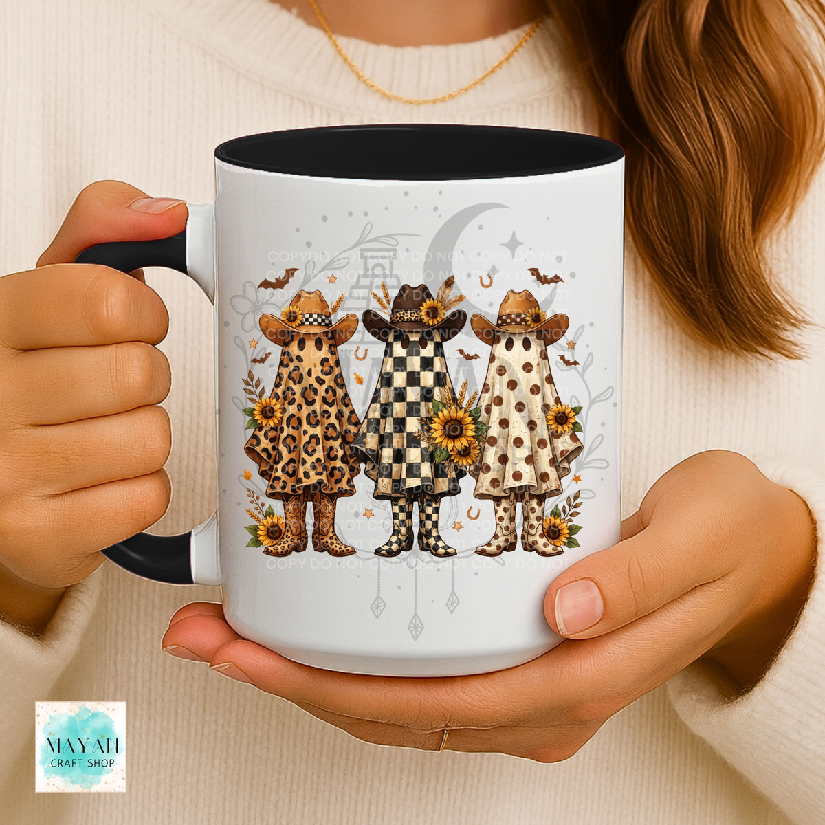 A person holds a white ceramic mug with a black interior and handle. The mug features a printed illustration of three witch characters in leopard, checkered, and polka-dot outfits with sunflowers and a crescent moon.
