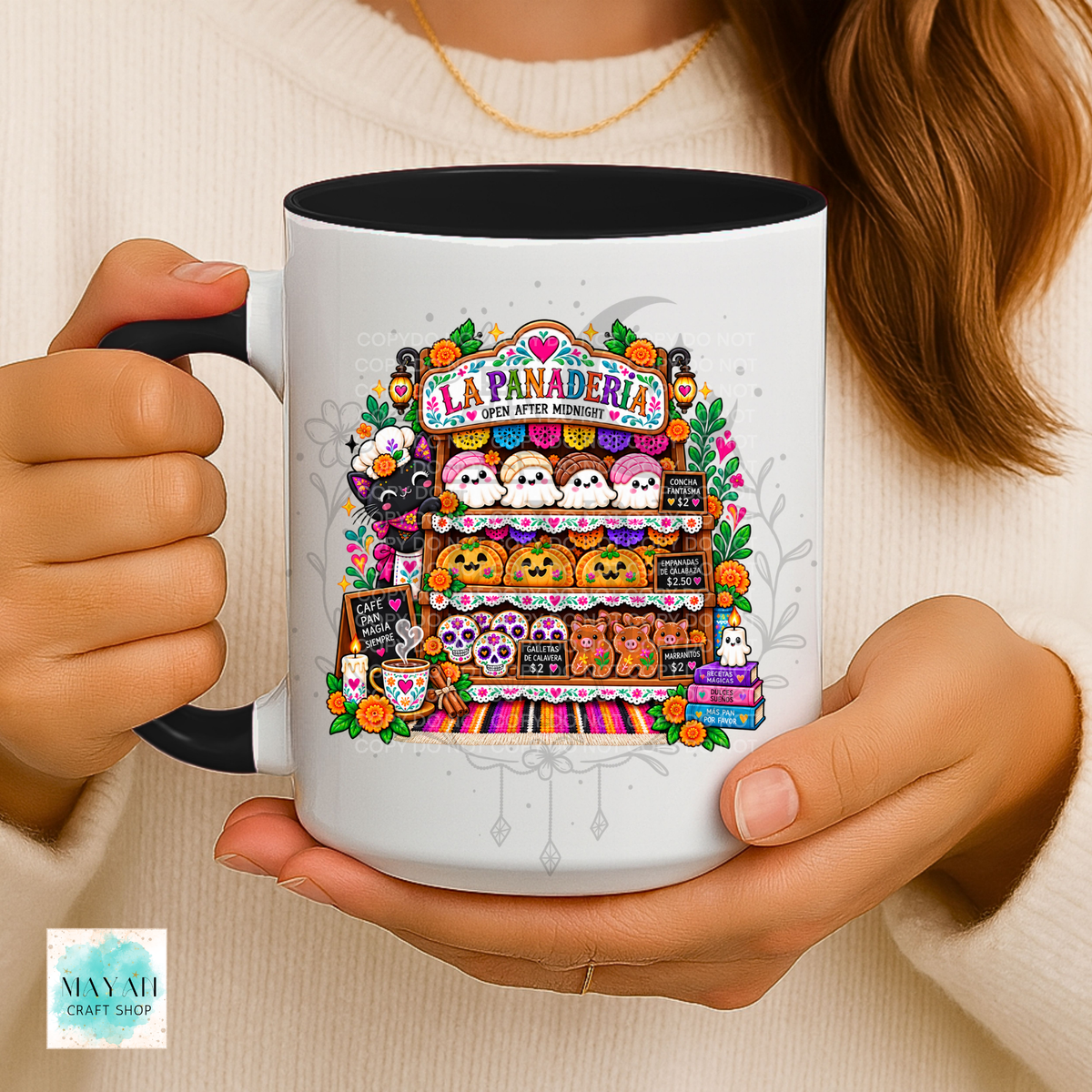A person holds a white ceramic mug with a black handle and interior. The mug features a colorful illustrated graphic of a Halloween-themed bakery display with pumpkins, ghosts, and the text 'LA PANADERIA'.