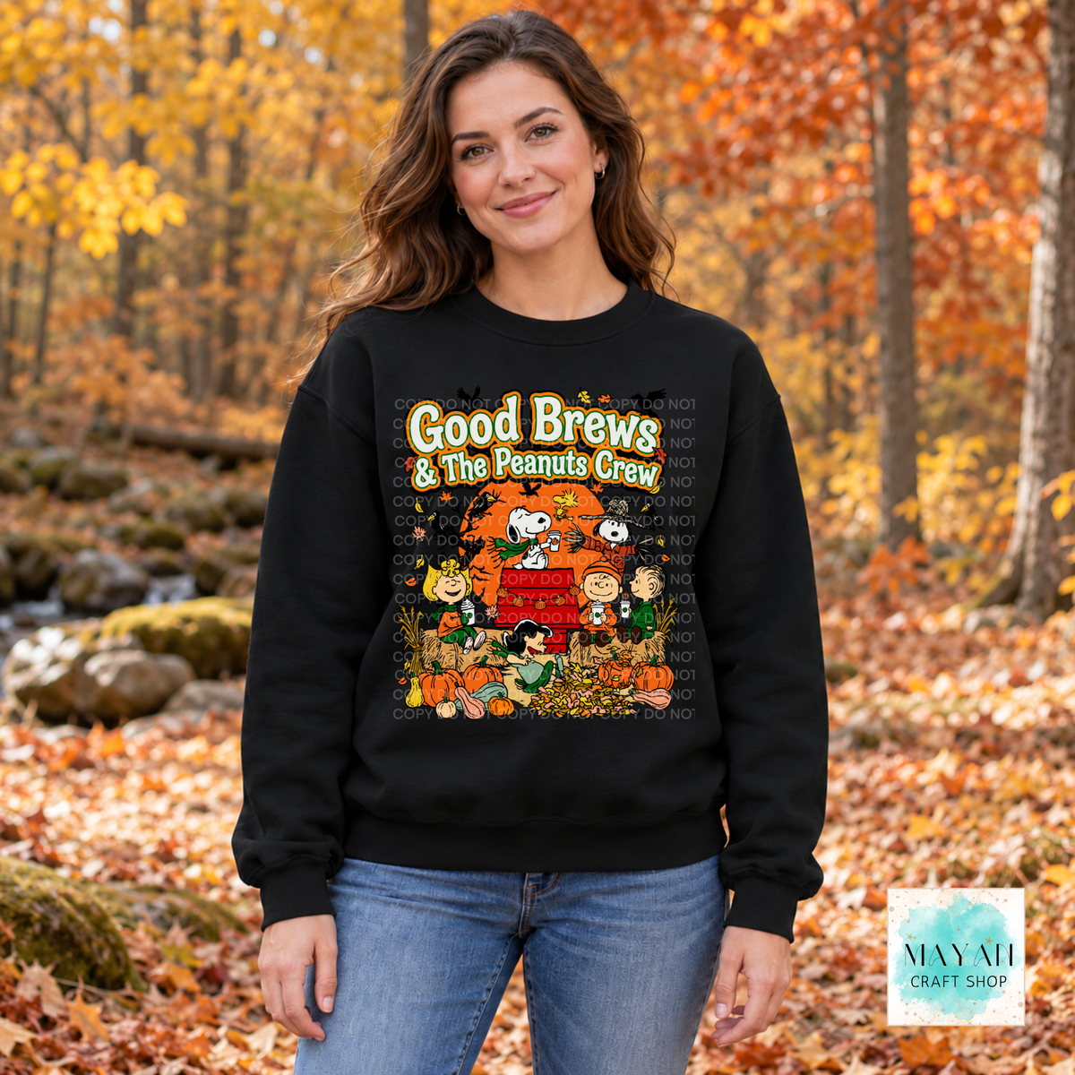 A woman wearing a black crewneck sweatshirt with a colorful 'Good Brews ' graphic, standing in an autumn forest with fallen leaves.