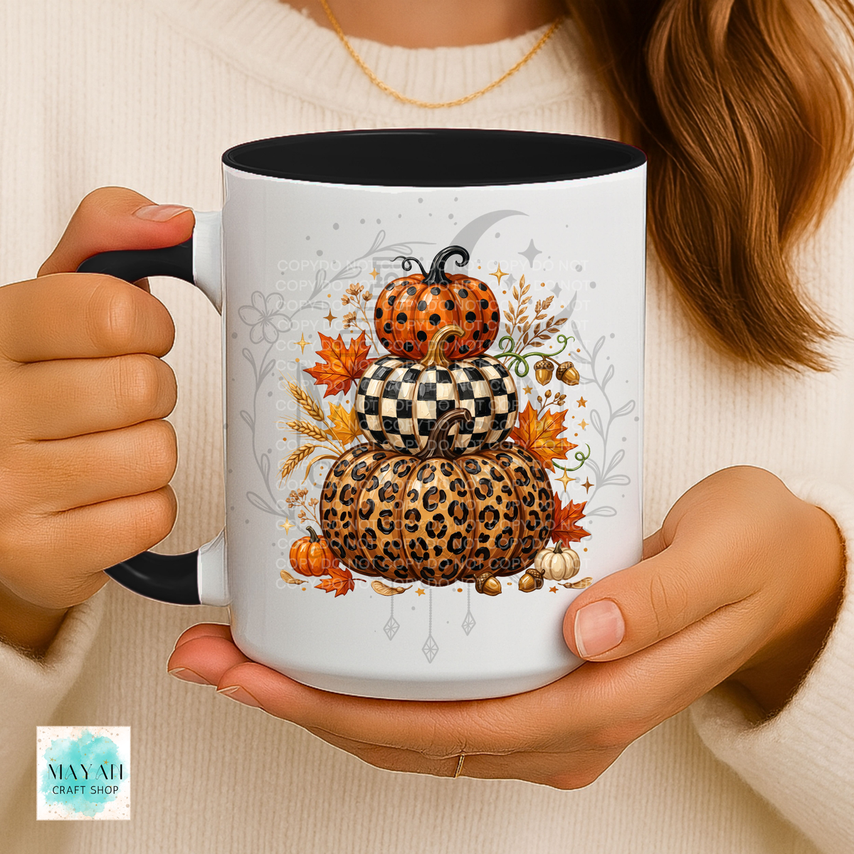 A person holds a white ceramic mug with a black handle and interior. The mug features a fall-themed graphic of stacked pumpkins in leopard print, checkerboard pattern, and orange with black spots, surrounded by autumn leaves and a crescent moon.